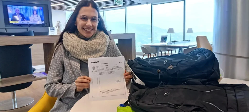 Mujer sonriente mostrando una guía electrónica de entrega de equipos frente a mochilas negras, en un ambiente de oficina moderna.