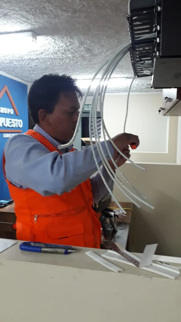 Técnico con chaleco naranja conectando cables blancos en un rack de comunicaciones dentro de una oficina.