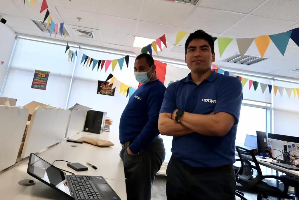 Dos técnicos de Okawa en oficina revisando laptops, uno sonriente mirando a cámara y otro trabajando en segundo plano.
