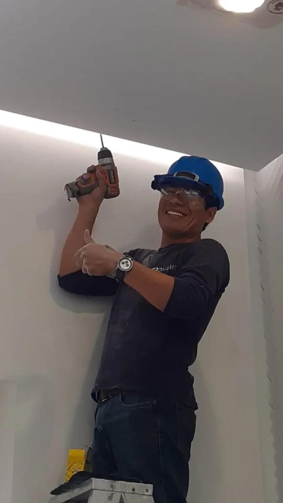 Técnico con casco y taladro instalando equipos en la pared, sonriente y levantando el pulgar.